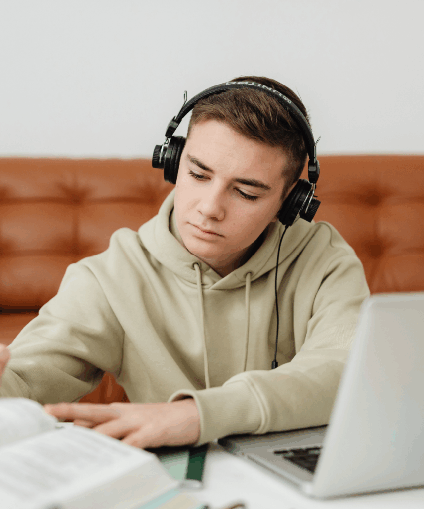 A person wearing headphones and a light-colored hoodie is studying at a desk with an open book and a laptop.