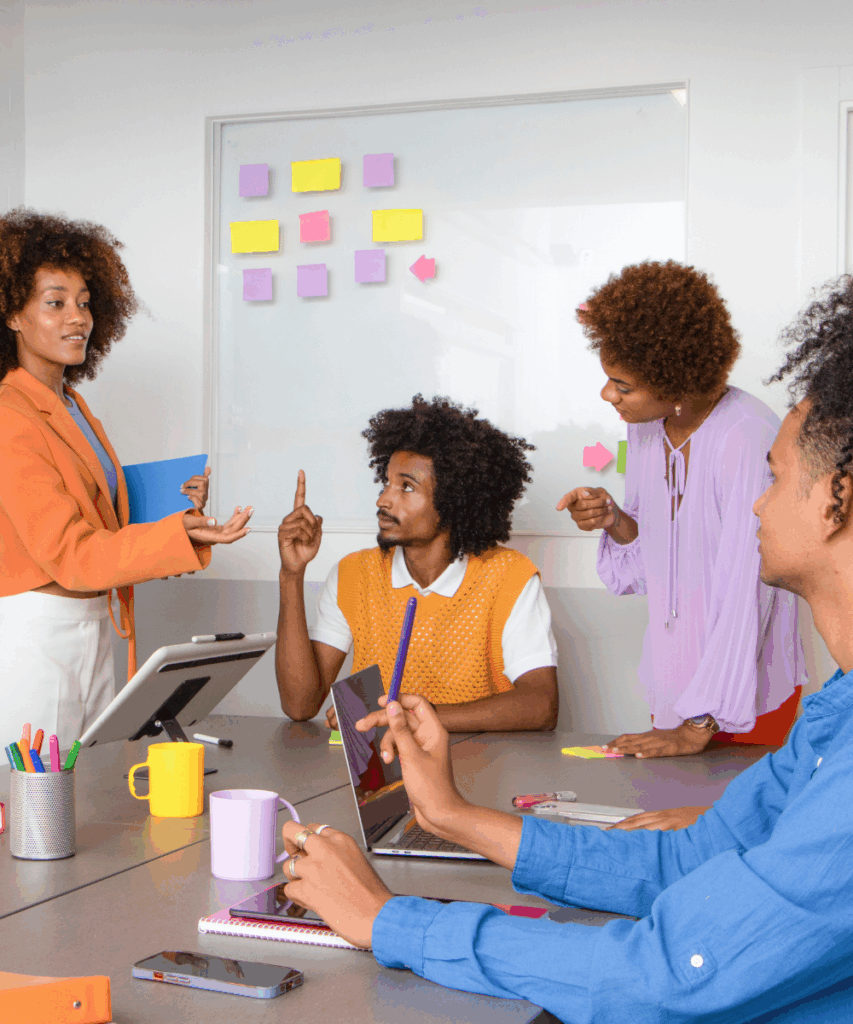 A group of coloured people engaging in a training conversation around a table in front of a white board with post stick notes.