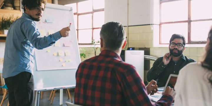 Creative team discussing ideas for startup project, speaker at board pointing at audience. Business colleagues in casual working together in contemporary office space. Brainstorming concept